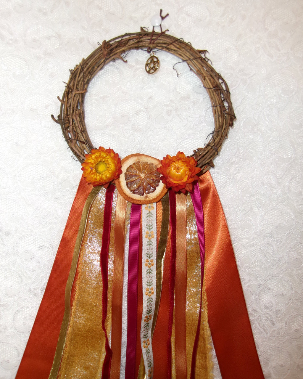 A rustic grapevine wreath decorated with orange dried flowers, a dried orange slice, and long flowing ribbons in orange, gold, pink, and white, laid flat on a white textured fabric background.