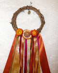A rustic grapevine wreath decorated with orange dried flowers, a dried orange slice, and long flowing ribbons in orange, gold, pink, and white, laid flat on a white textured fabric background.